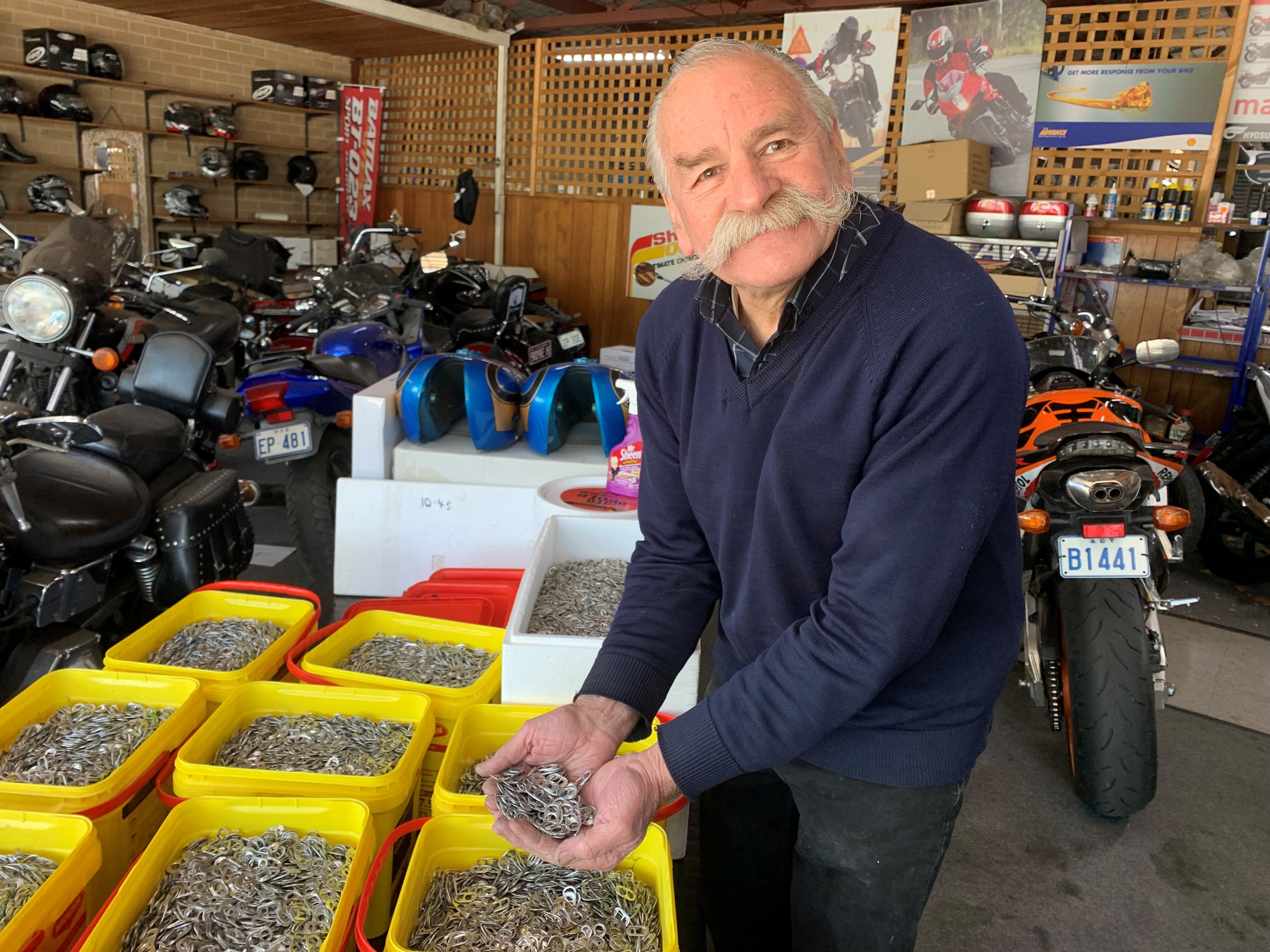 An older man with his hands full of drink can ring pulls, surrounded by buckets of ringpulls.