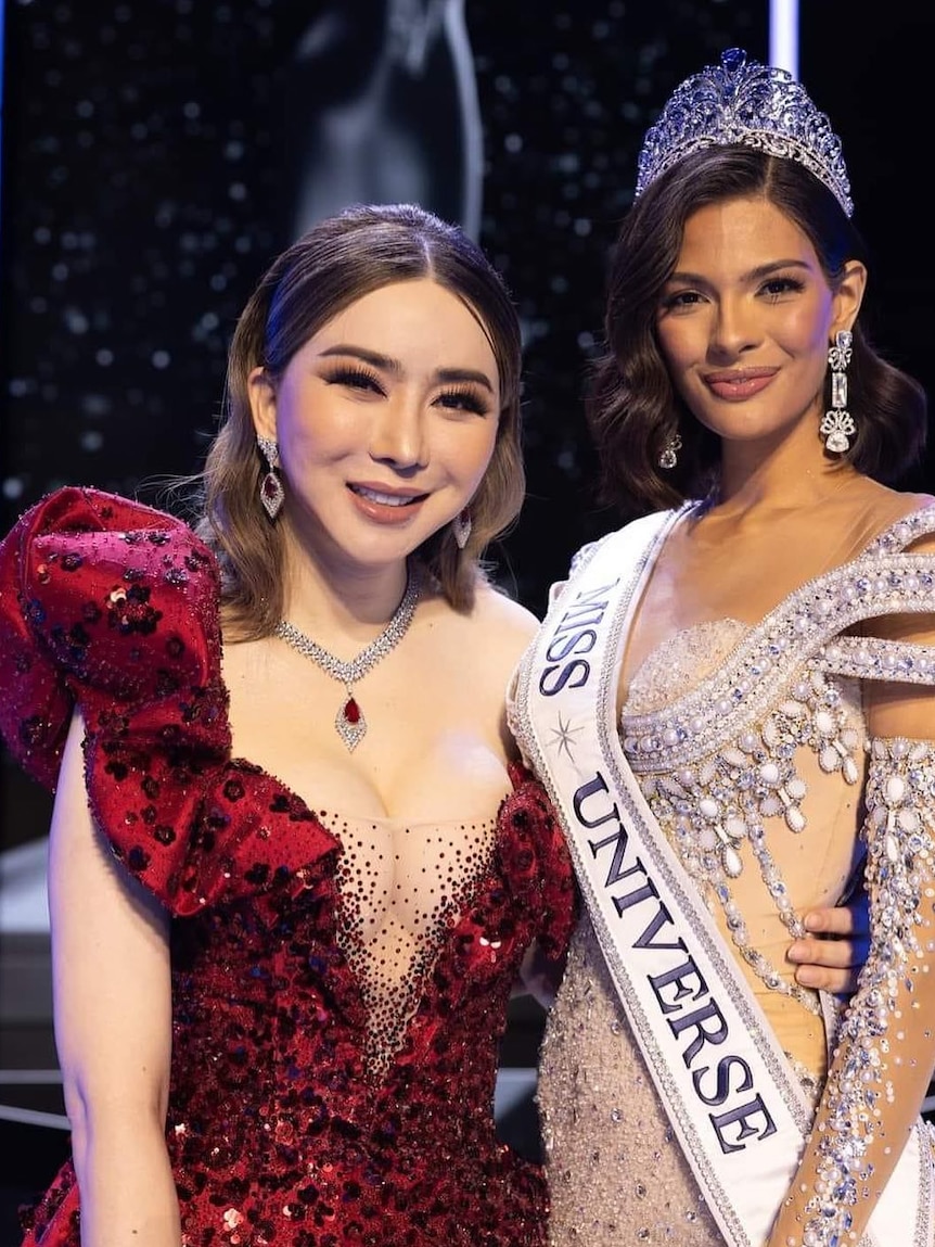 Two women in evening gowns, one wearing a giant bedazzled crown and MISS UNIVERSE sash, smile together