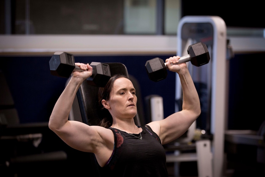 Mandy Hagstrom performs an inclined shoulder press with barbells at a gym.