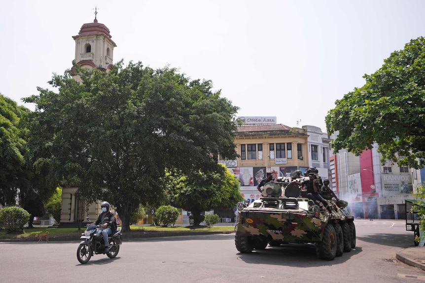 A camouflaged army tank drives through a town square passing a motorbike with soldiers sitting on its exterior.
