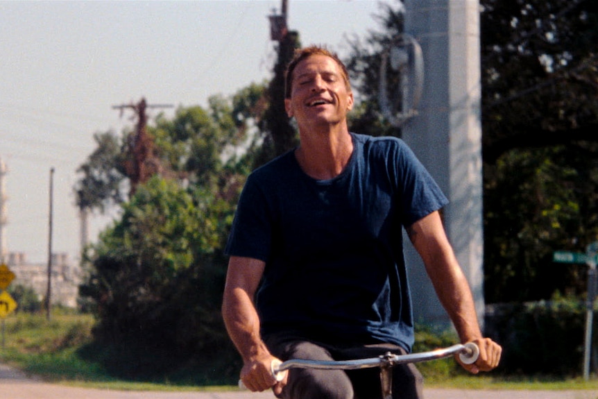 A 40-something man in a navy shirt grins as he rides a bicycle down an empty street in an industrial town