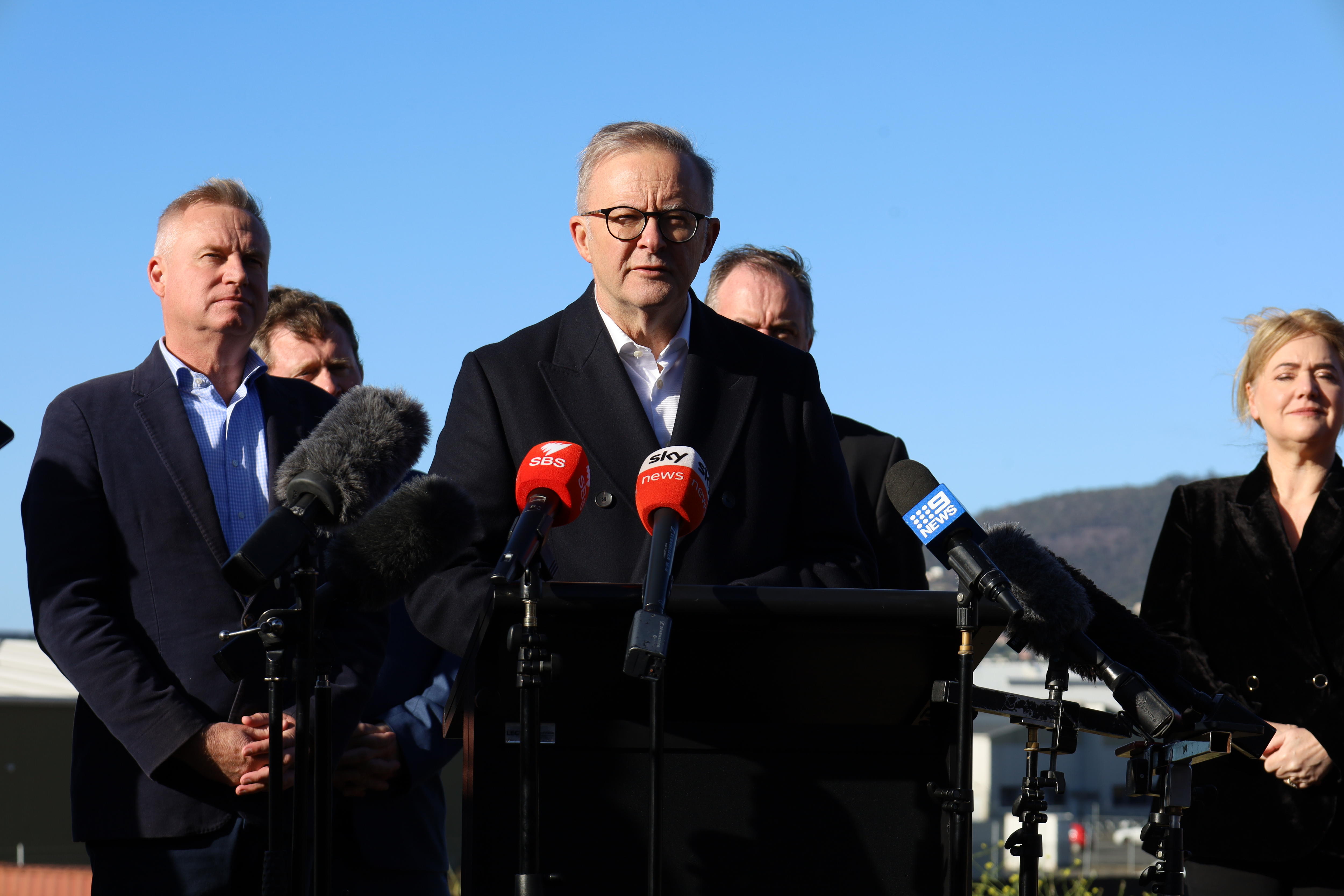 A balding man wearing glasses stands in front of other people at a lectern with microphones