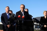 A balding man wearing glasses stands in front of other people at a lectern with microphones