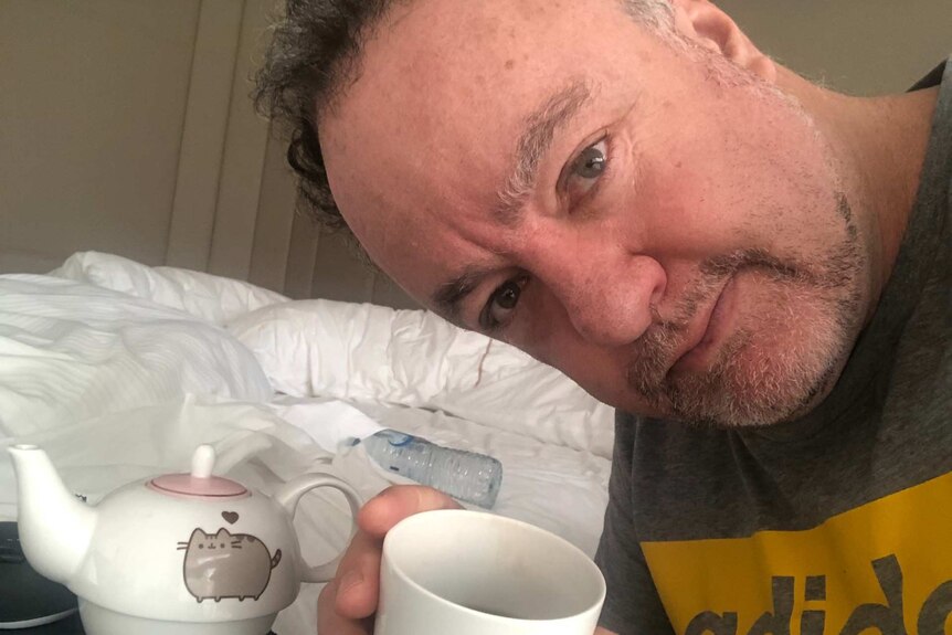 James take a selfie holding a mug in a hotel room.