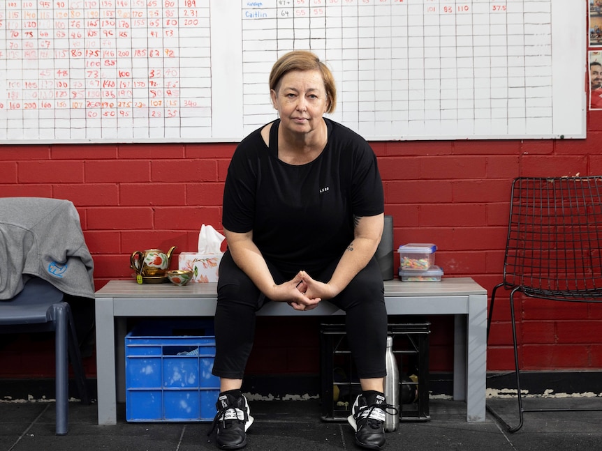 A middle aged woman sits on a table in a gym, in front of a whiteboard