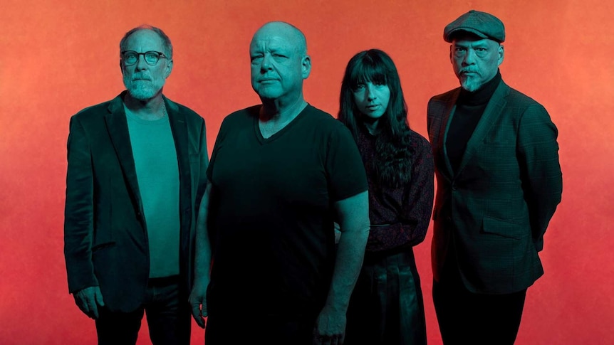 Alt-rock musicians Pixies are standing next to each other looking at the camera seriously in front of an orange wall.