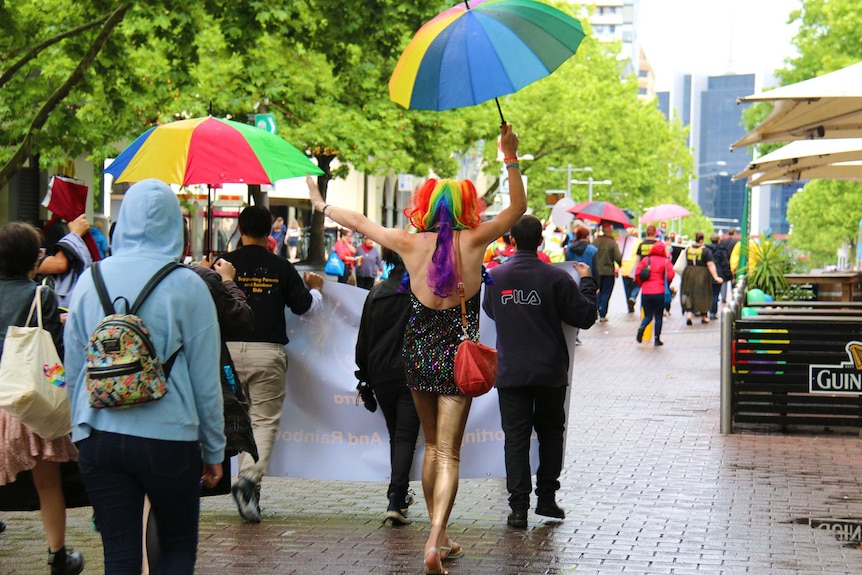 A person with rainbow hair, holding a rainbow umbrella and wearing a sparkly dress and leggings dances down the street.