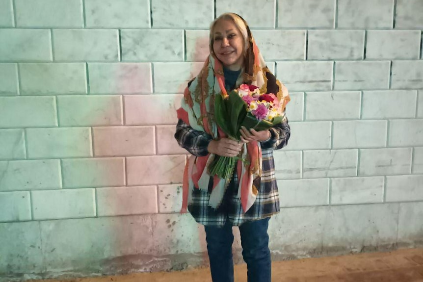 A woman wearing a headdress stands in front of a white brick wall, holding a bouquet of flowers.