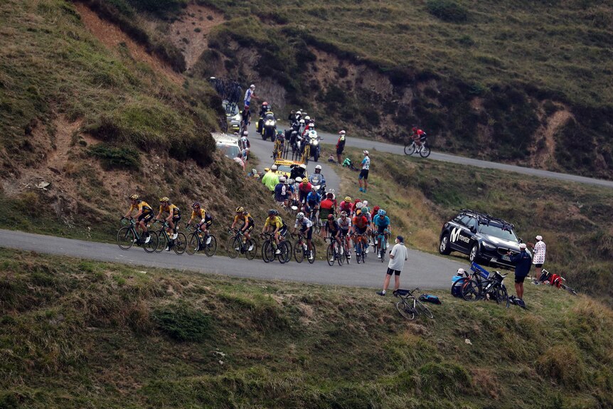 Riders cycle up a mountain in a line with a couple of people standing watching