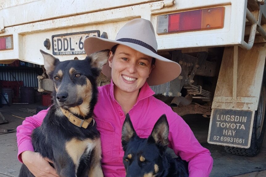A woman sits on the ground behind a ute cuddling two dogs.