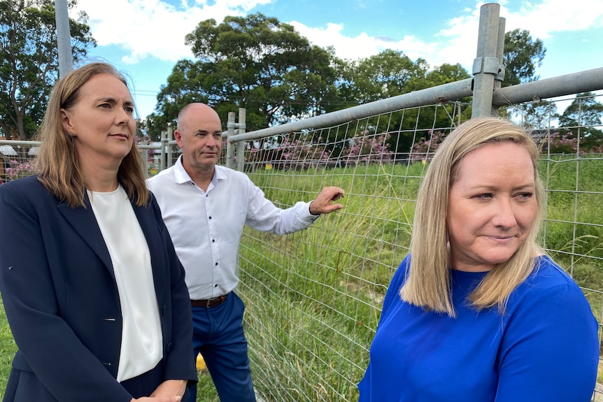 Two women and a man look pensive as they stand by fence and empty field. 