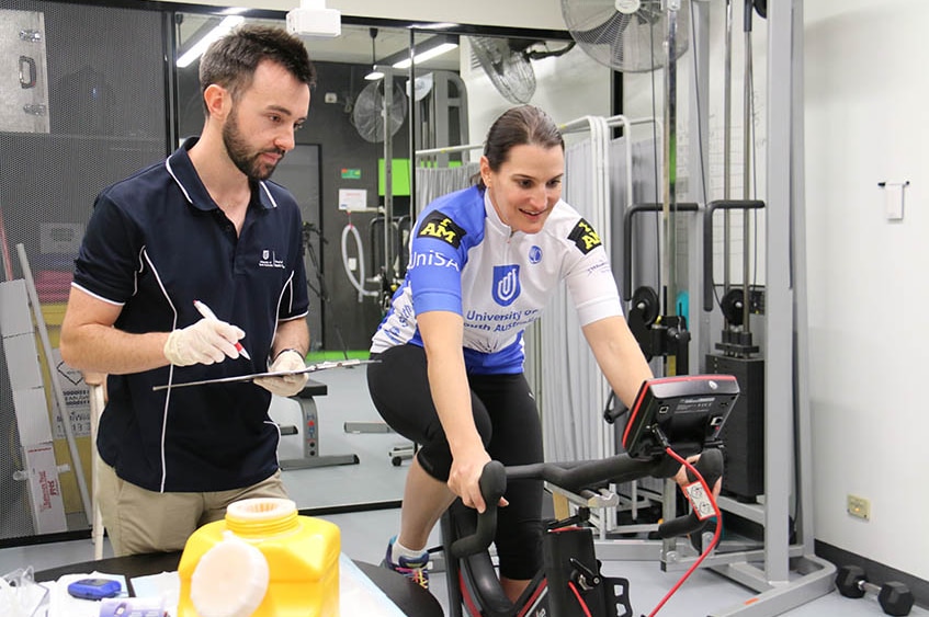 A woman races on a cycling machine while a man looks over taking observations