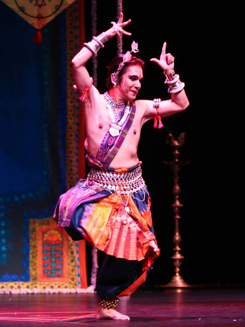 A man in traditional Indian costume, makeup and jewelry performs a dance move on stage.