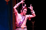 A man in traditional Indian costume, makeup and jewelry performs a dance move on stage.