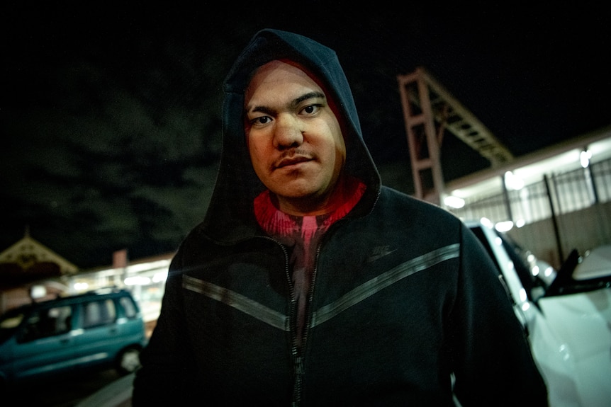 A man wearing a hood looks down at the camera while standing in a carpark at night.
