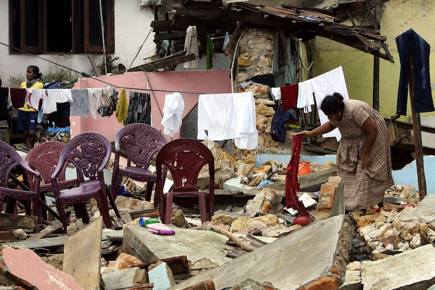 A woman bends down to pick up debris in the rubble of a destroyed building. Washing still hangs on the line