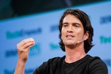 Adam Neumann gestures with one hand while speaking with a microphone next to his mouth. He appears in front of a blue background