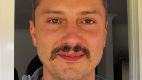 A man with a moustache smiles at the camera.
