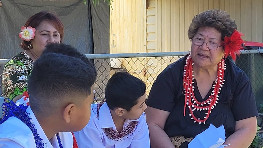 Mele is wearing a red flower and red necklace. She's pictured sitting with two boys and another woman. 