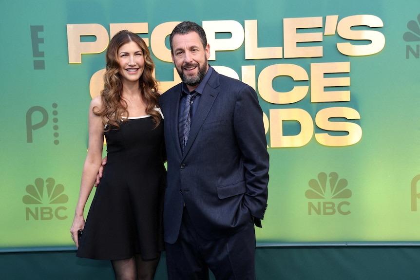 Adam Sander dressed in an all black suit and wife in black dress standing in front of a green wall with lettering