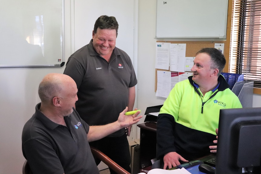 Three men sit in an office facing each other having a laugh.