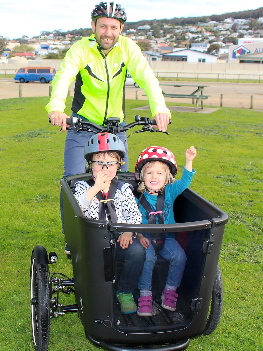 Two children sitting in a cargo carrier on an electric bicycle with a man on the saddle and carpark.