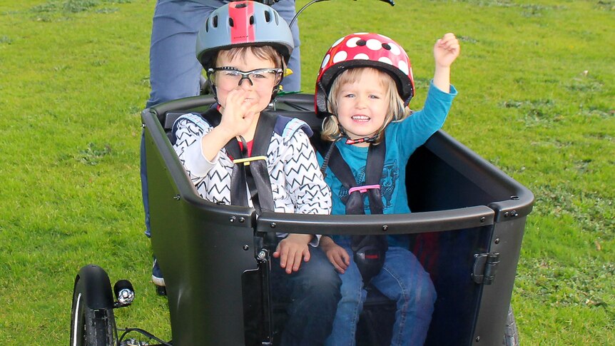 Two children sitting in a cargo carrier on an electric bicycle with a man on the saddle and carpark.