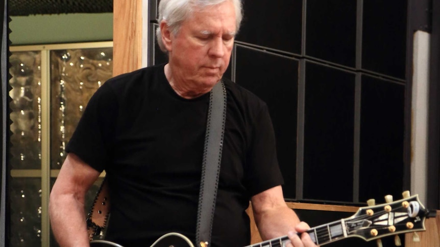 James Williamson wearing black trousers and a black tee shirt playing a black guitar in a music studio