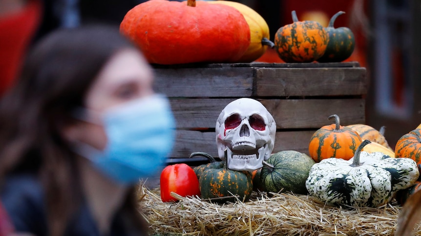 A woman in a disposable face masks walks in front of a fake skull and pumpkins laid out for Halloween.