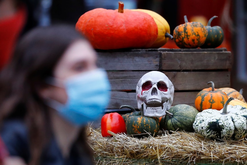 A woman in a disposable face mask walks in front of a fake skull and pumpkin laid out for Halloween.