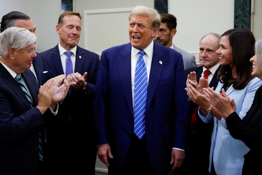 Donald Trump is standing while surrounded by a group of politicians, who are applauding.