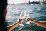 two glasses of white wine coming together as a cheer with a background water in an ocean