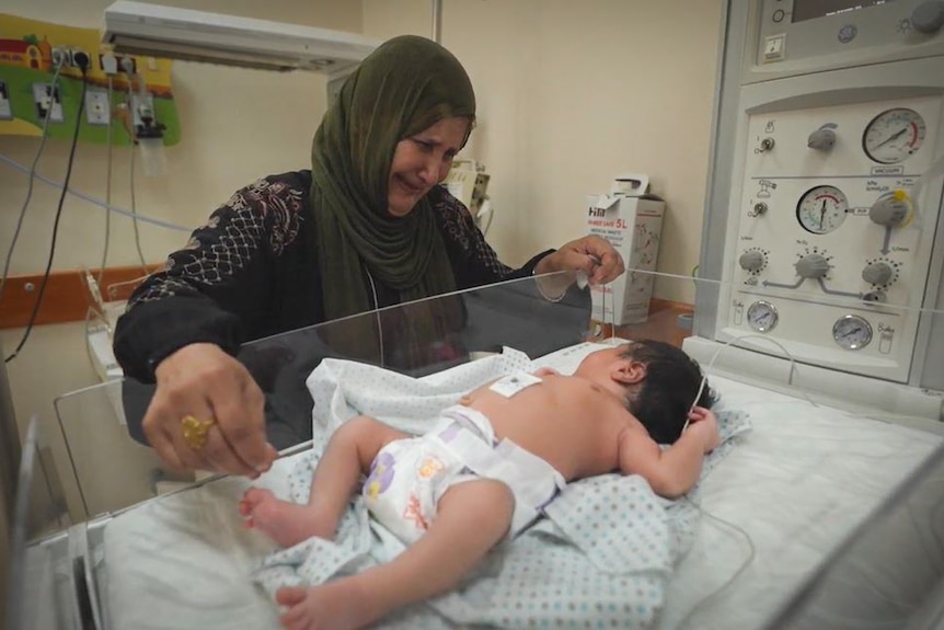 一名戴头巾的女性依靠在病床边看着一名婴儿