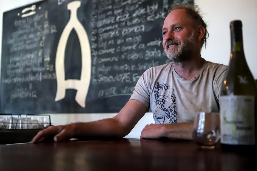 Man with off white t-shirt sits at wooden table with chalkboard in background and wine glass and bottle in foreground