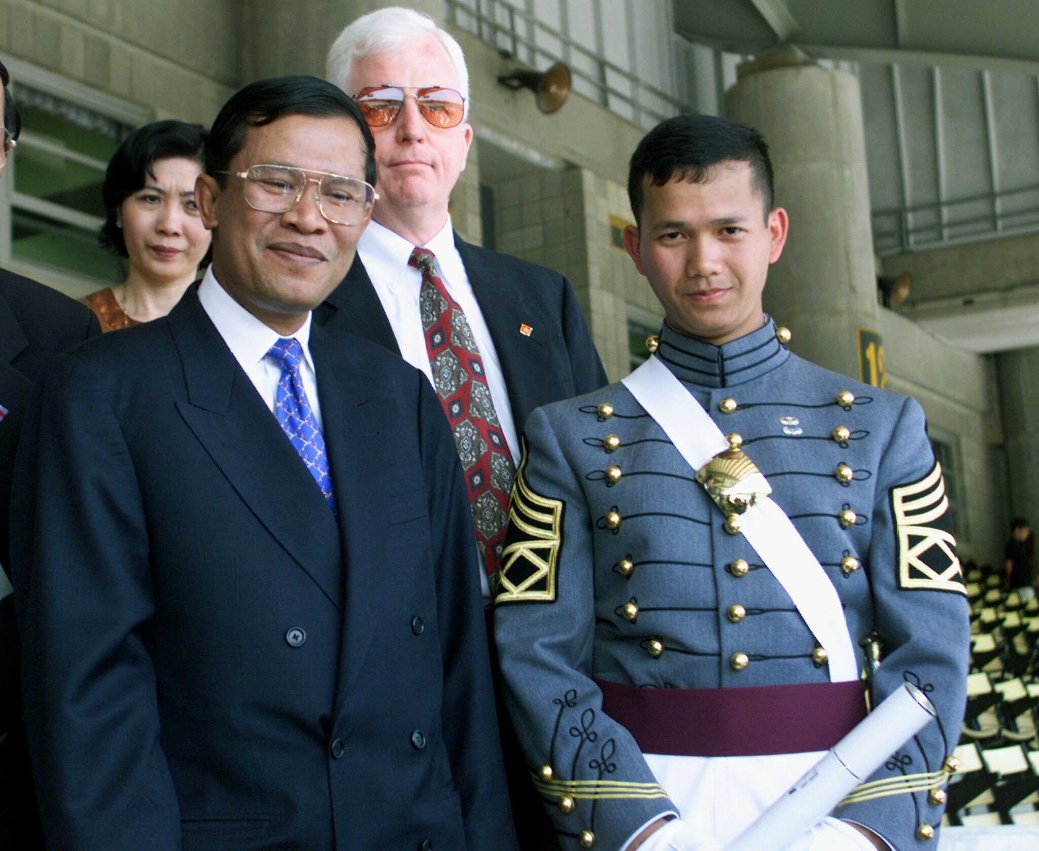 An older man in navy suit with bright blue tie stands next to young man in formal military uniform