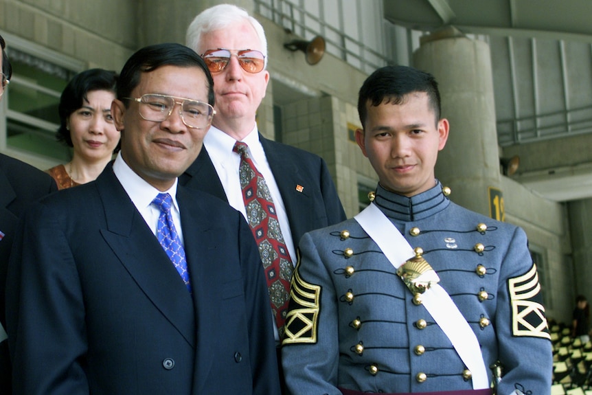 An older man in navy suit with bright blue tie stands next to young man in formal military uniform