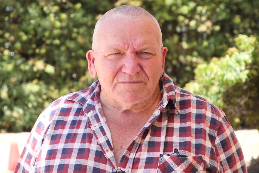 A head and shoulders mid-shot of an older man wearing a red-white and blue checked shirt outside.