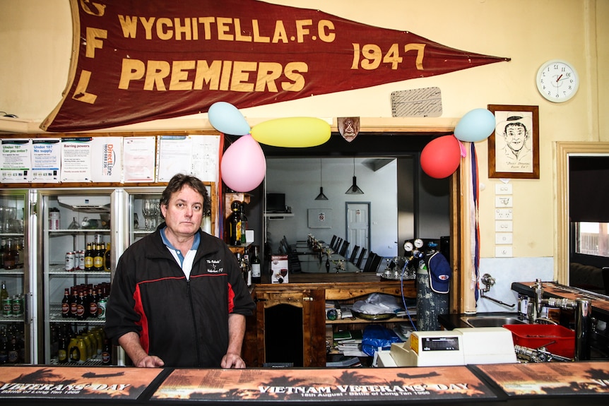 A man standing in front of hotel fridges, with a 1947 premiership banner hanging on the wall.