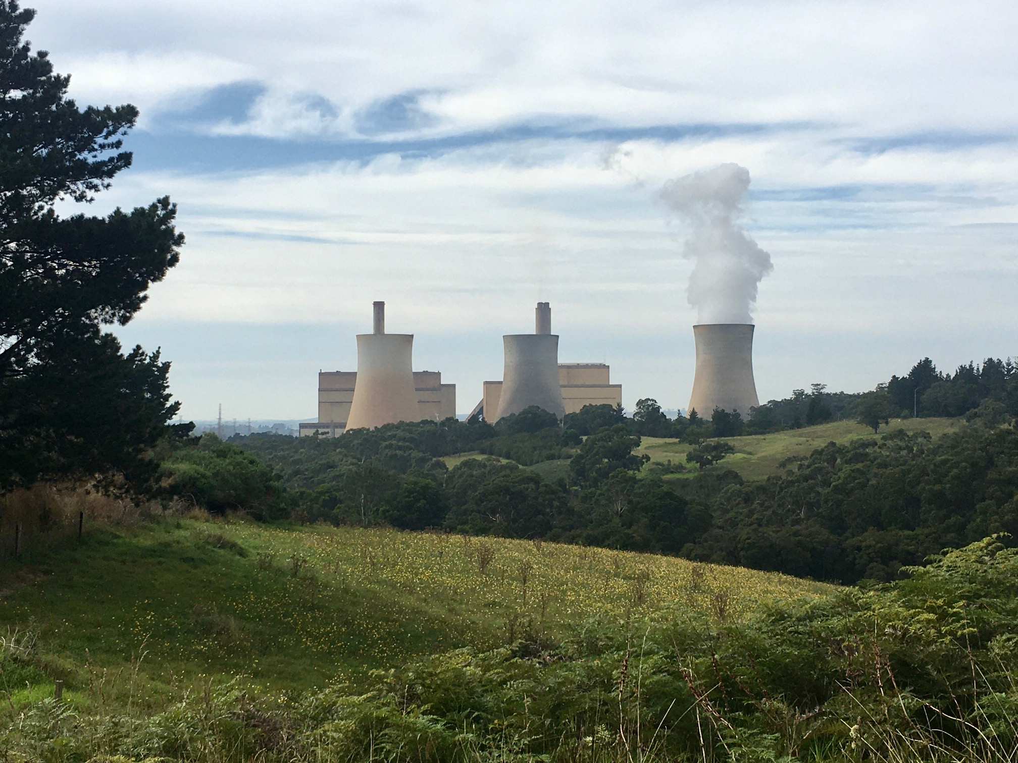 A large power plant blowing smoke with green rolling hills in the foreground.