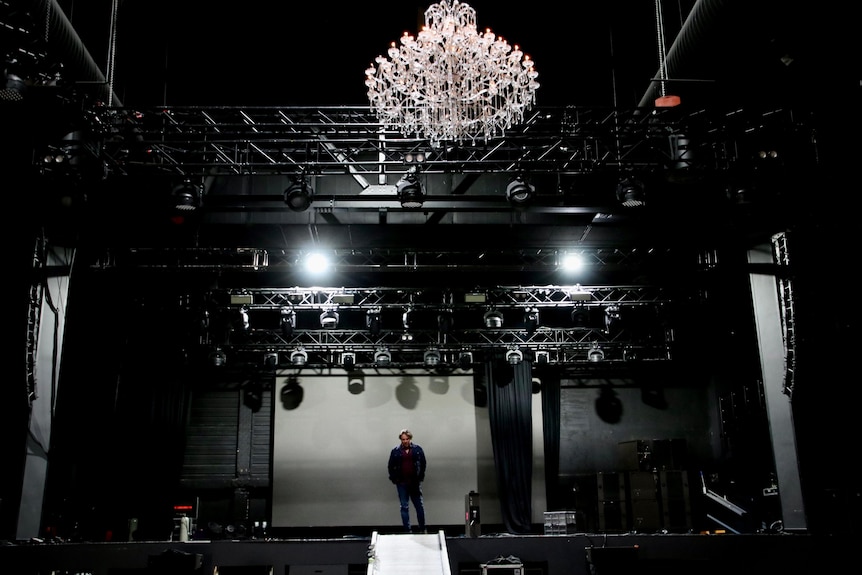 JC se tient sur une scène sombre, avec un grand lustre au-dessus 
