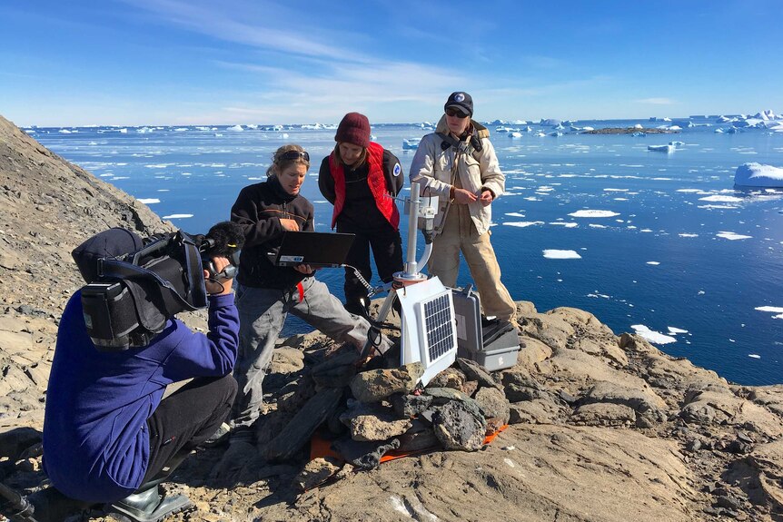 Three people are filmed operating equipment on a island with icebergs in the background.