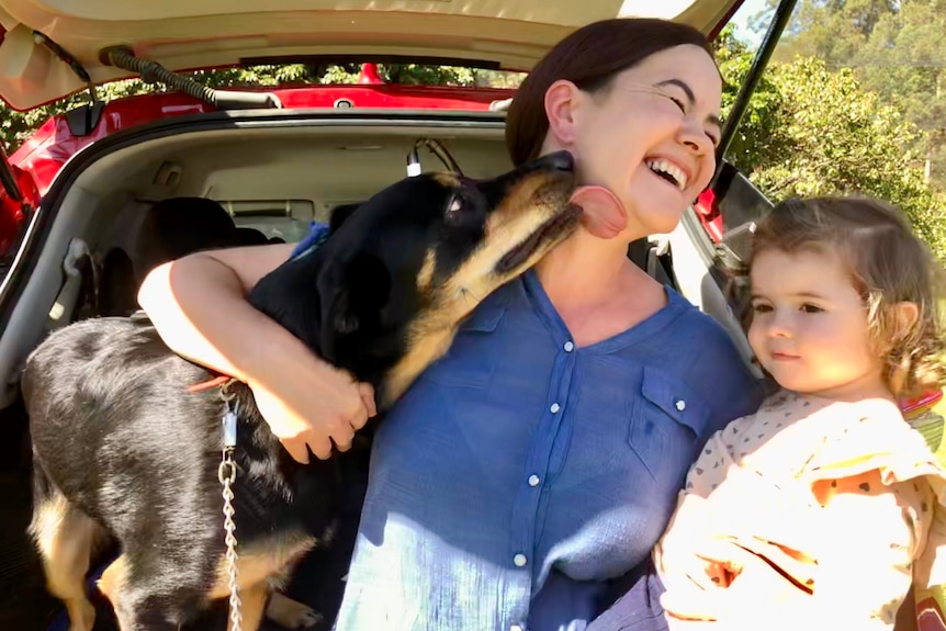 Dog licks ladies face as she holds her daughter. 
