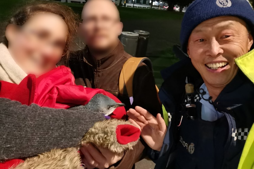 Two people with faces blurred and a police officer pose with a blue penguin