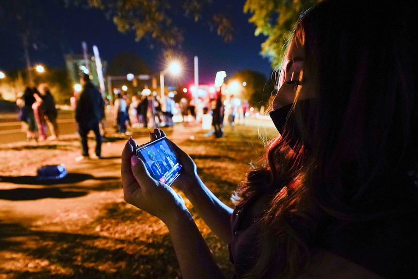 A woman with brown hair wearing a mask looks down at a phone displaying the debate while standing at a park.