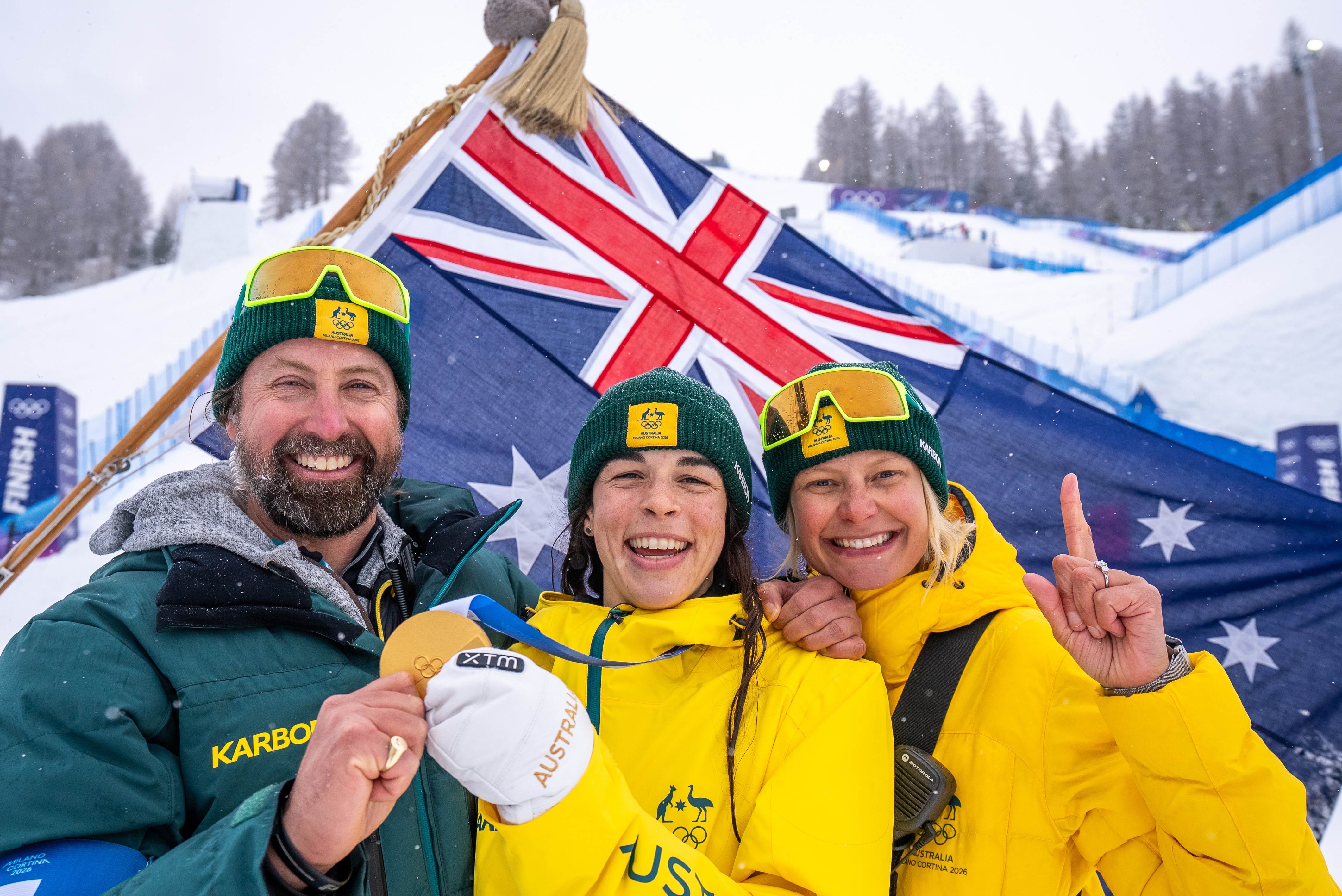 A smiling man and two women, one holding a gold medal, in front of an Australian flag on a snowfield.