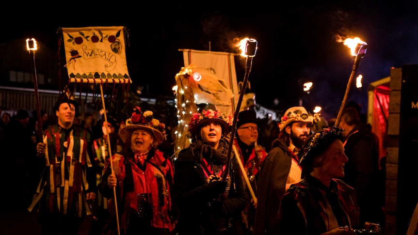 A group of people wearing robes, flower crowns carry fire-lit torches in the dark.
