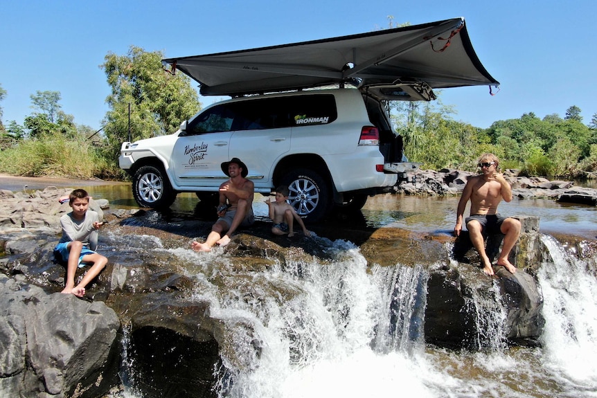 Two men and two children sitting on river crossing with four wheel drive behind them. Blue skies and Green bushland.