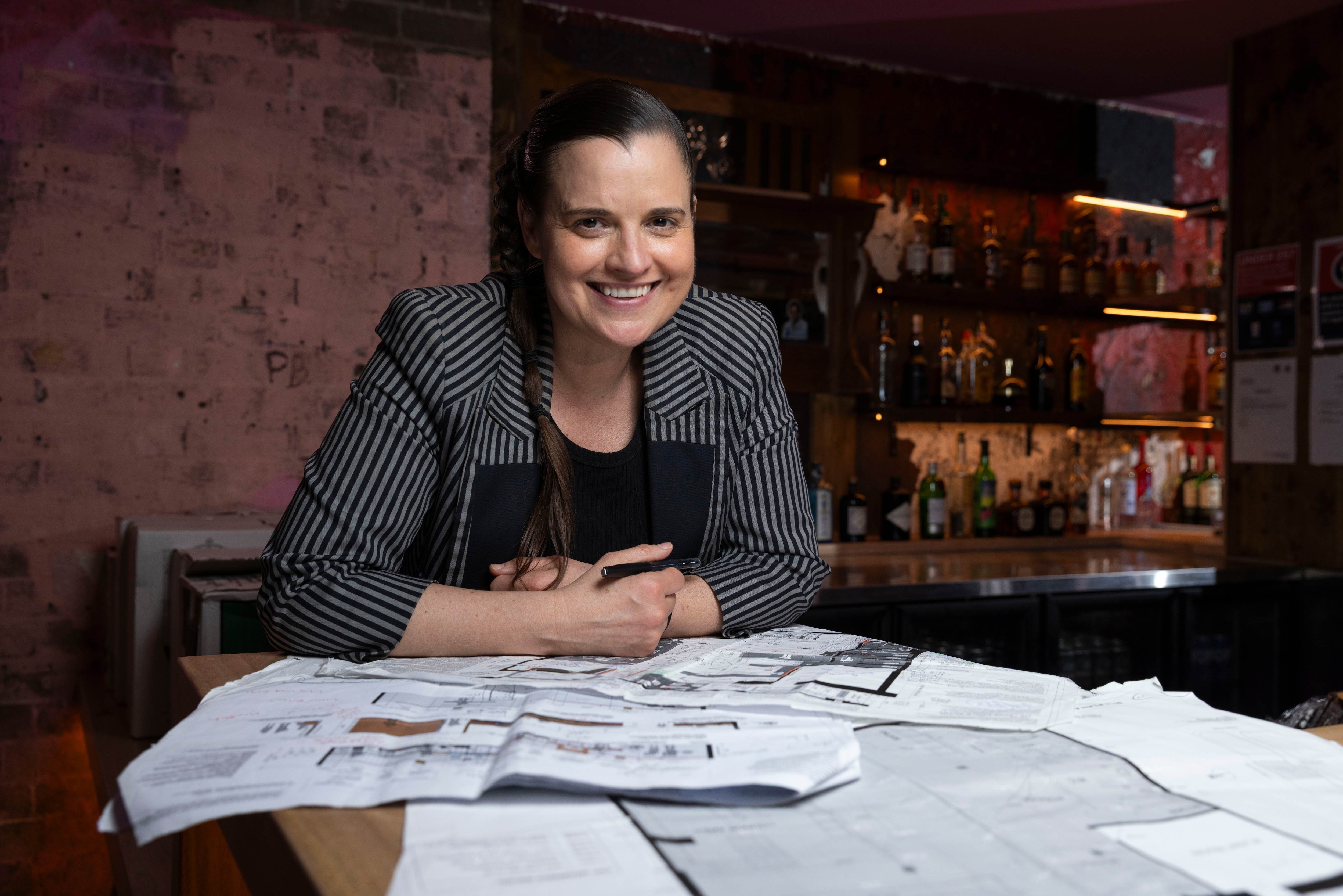 Kristie Paul with a brunette braid, a striped blazer, smiling near a bar and table full of blueprints.
