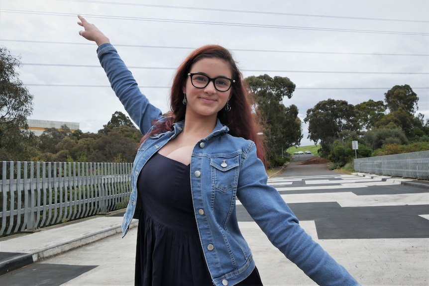 A girl with dark red hair and glasses wearing a denim jacket holding a dance position with her right arm in the air