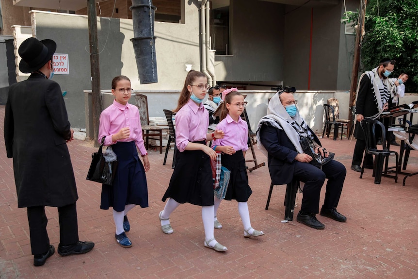 Three girls in pink and blue uniforms with masks around their necks walk next to three men praying.